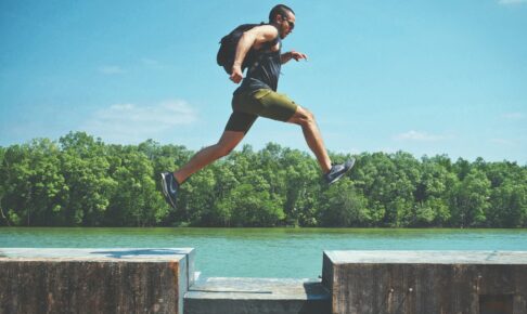 man leaping on concrete surface near body of water and forest at the distance during day