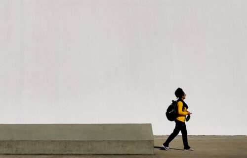 human in yellow long-sleeved T-shirt walking beside white concrete wall