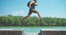 man leaping on concrete surface near body of water and forest at the distance during day